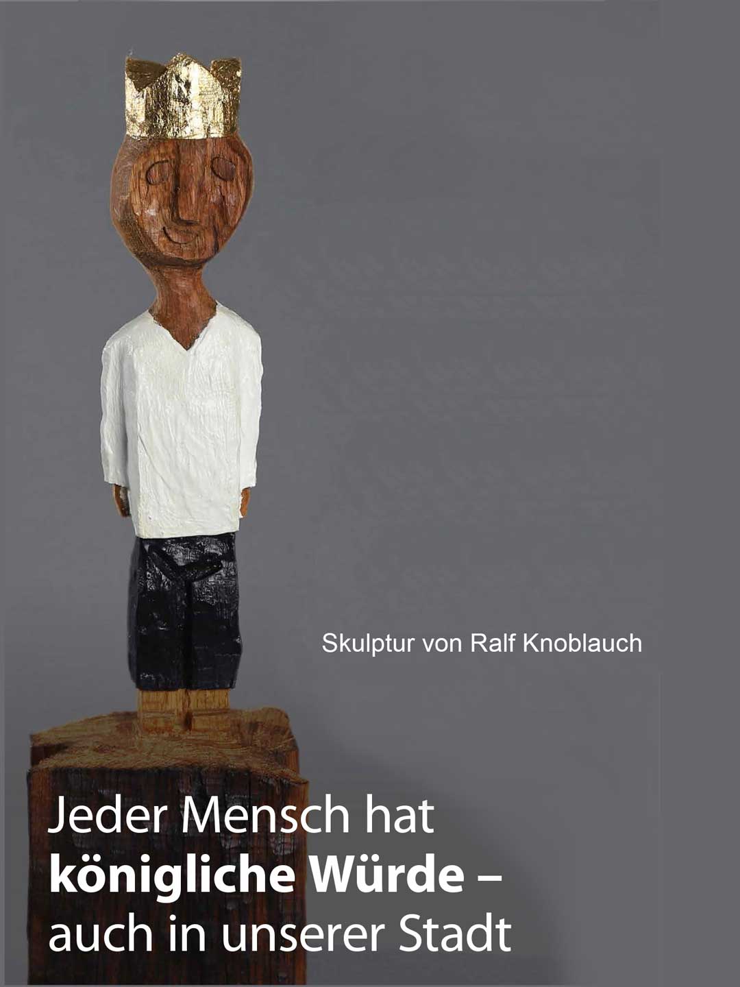 Skulptur von Ralf Knoblauch - Dargestellt ist eine männliche Figur aus Holz geschnitzt mit weißen Hemd, schwarzer Hose und einer Golgkrone auf dem Kopf